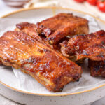 saucy cooked ribs on plate with vine tomatoes behind.