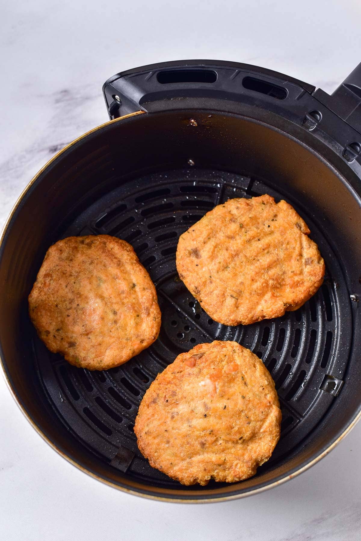 three cooked salmon burger patties in round black air fryer basket.