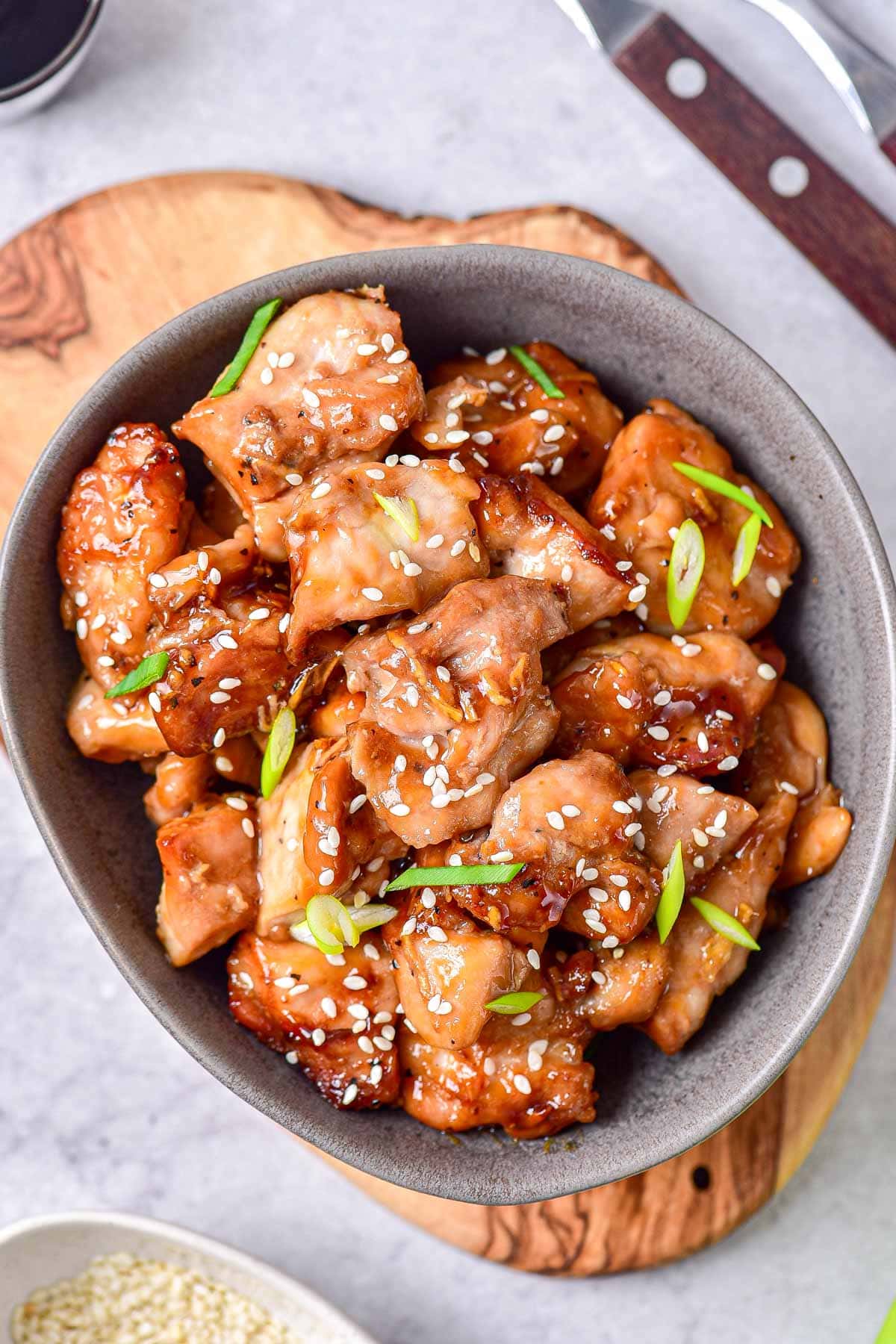 grey bowl on wooden board with pieces of teriyaki chicken topped with sesame seeds and green onion.