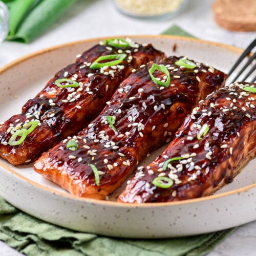 three teriyaki salmon on plate with sesame seeds on top and silver fork beside.