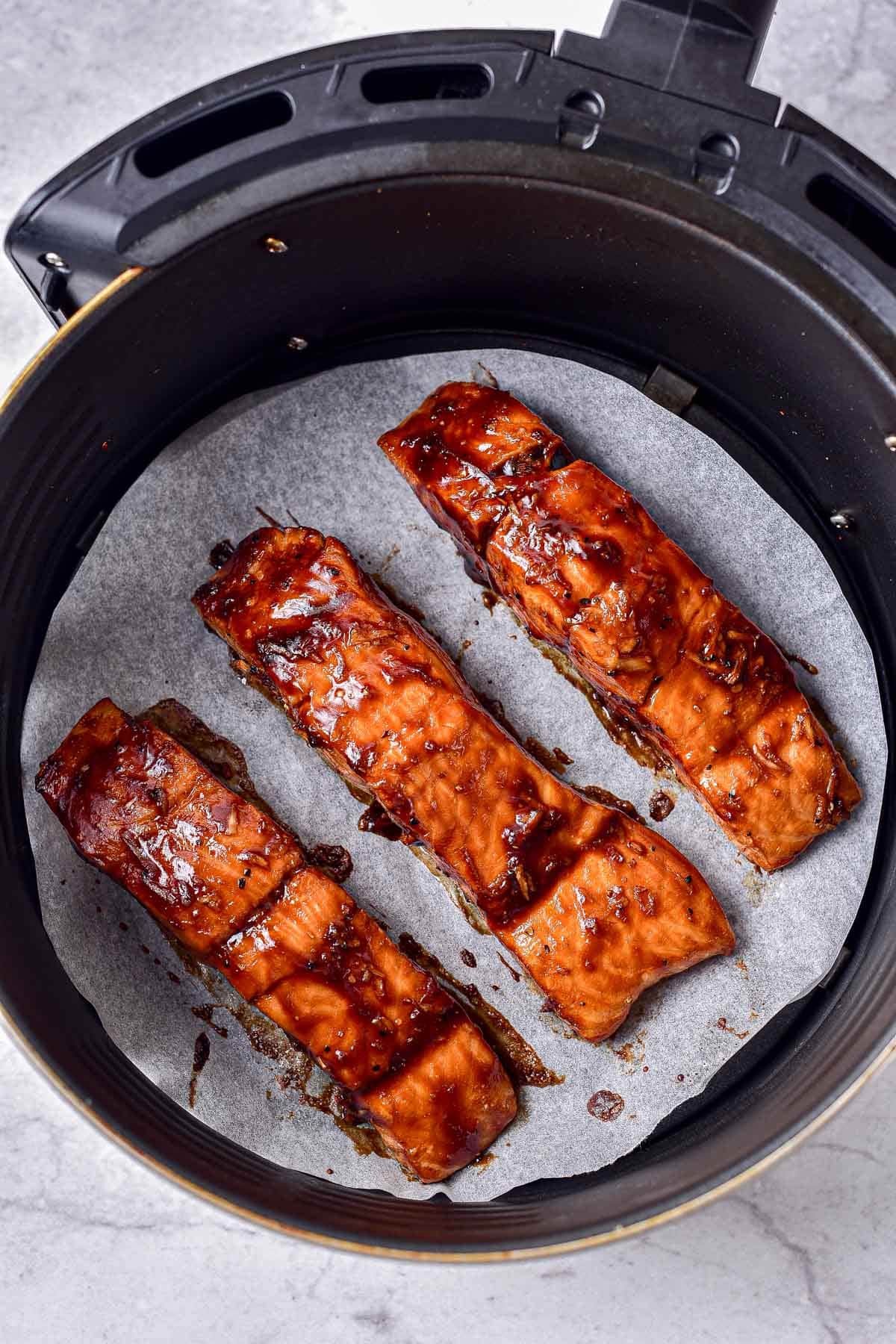 cooked teriyaki salmon on parchment paper in air fryer basket on counter.