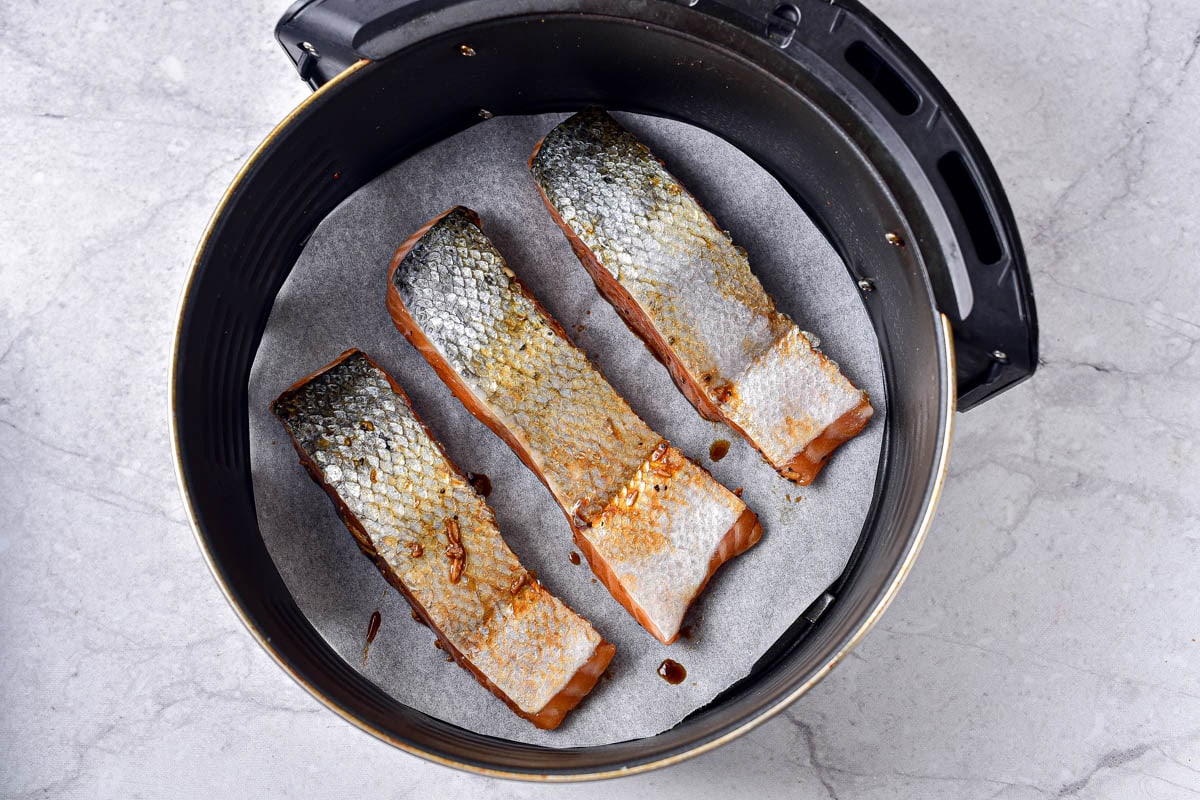 three pieces of marinaded salmon sitting on parchment paper in round air fryer basket.