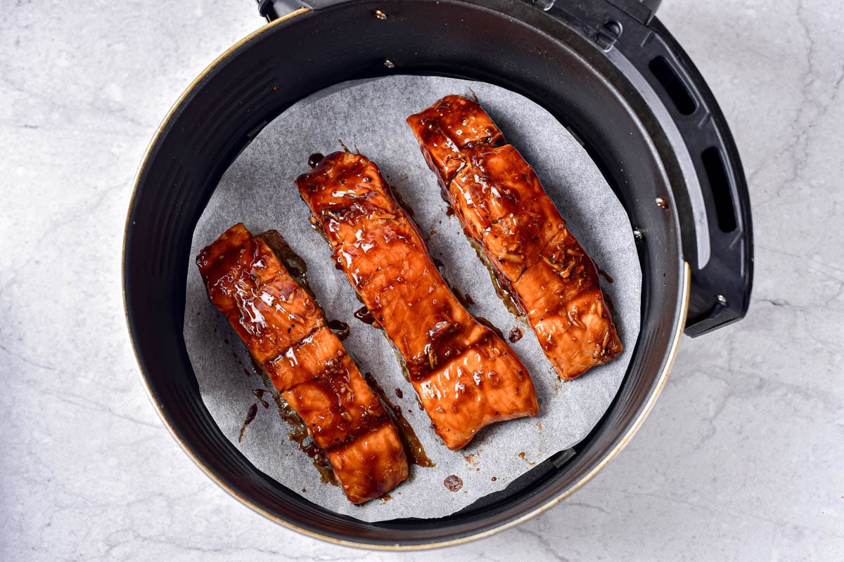 cooked teriyaki salmon on parchment paper in round black air fryer basket.