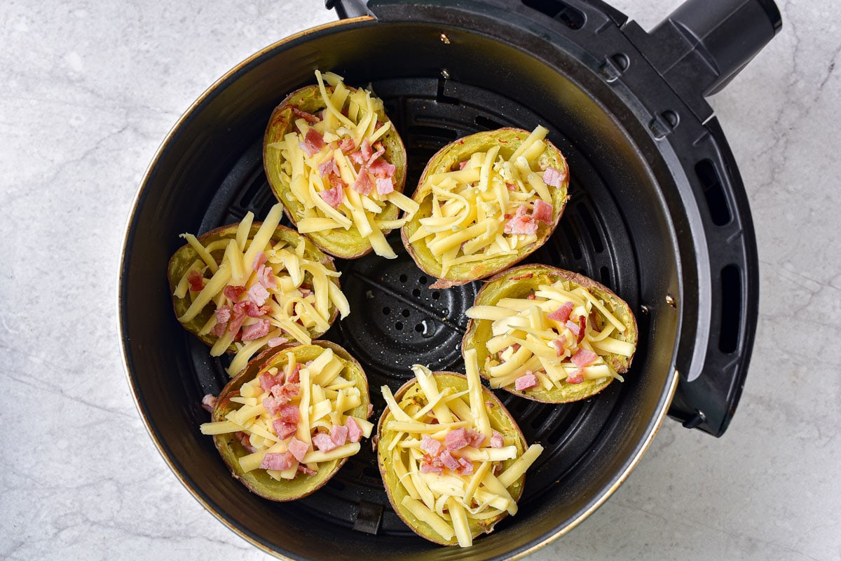 potato skins in round air fryer basket with shredded cheese and bacon on top.