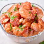 glass bowl filled with bacon wrapped shrimp covered in green herbs.