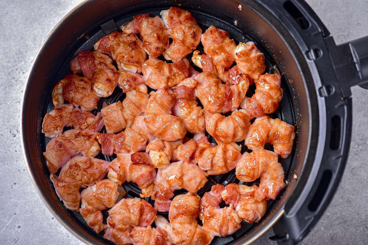 partially cooked shrimp wrapped in bacon in black air fryer basket on counter.