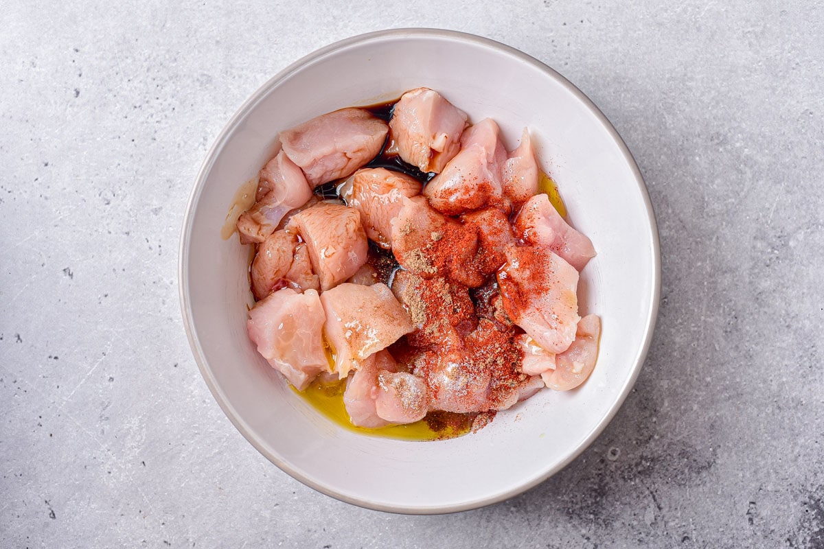 chunks of cut chicken in white bowl on counter with spices on top.