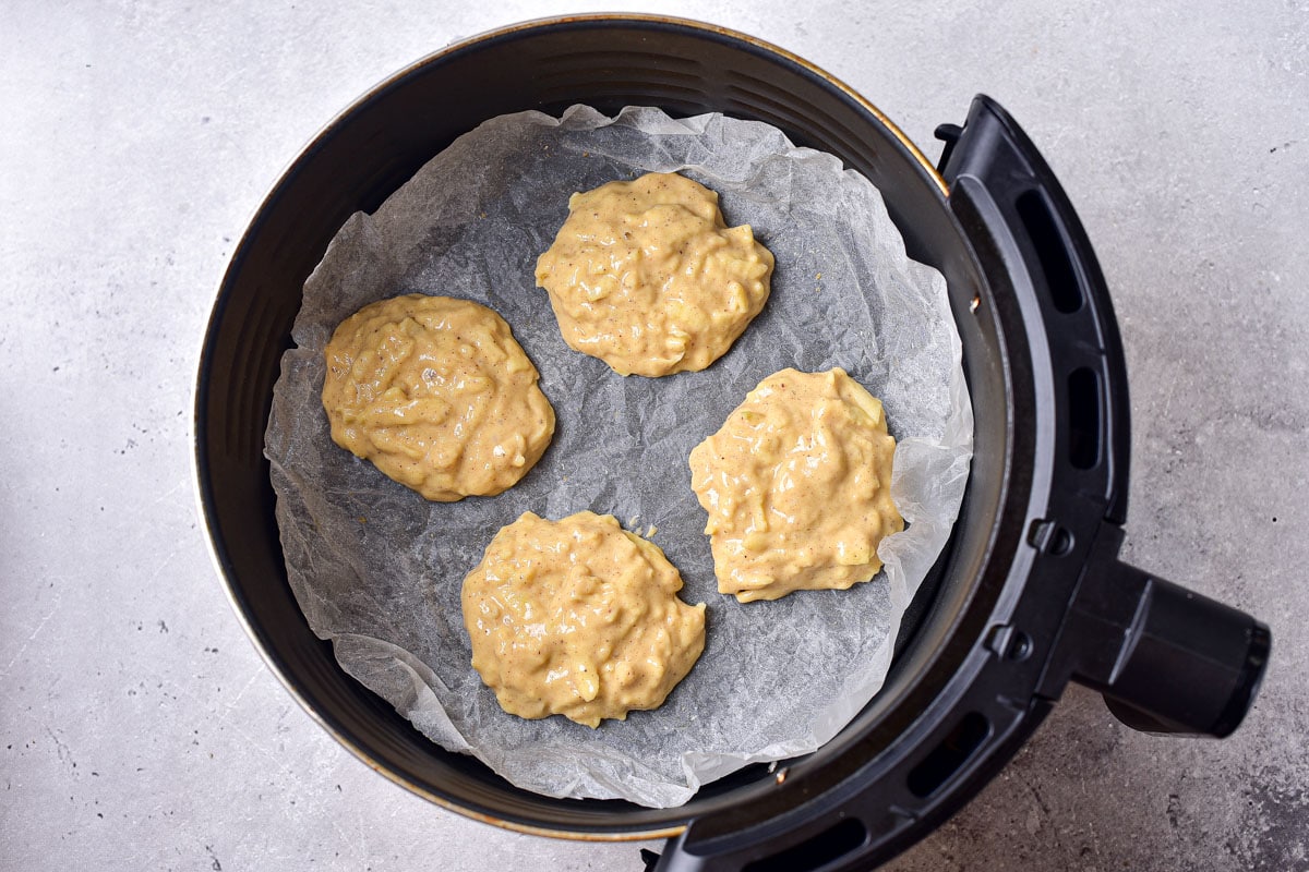 four piles of apple fritter batter on parchment paper in round air fryer tray.