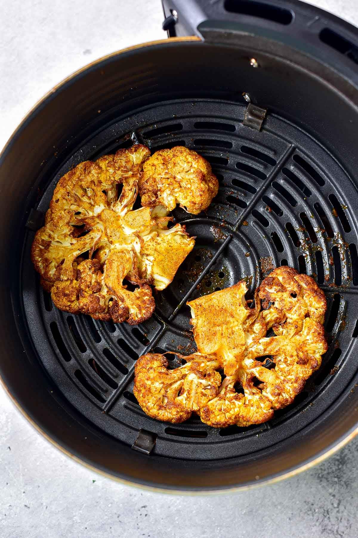 two cooked cauliflower steaks in round black air fryer tray on counter.