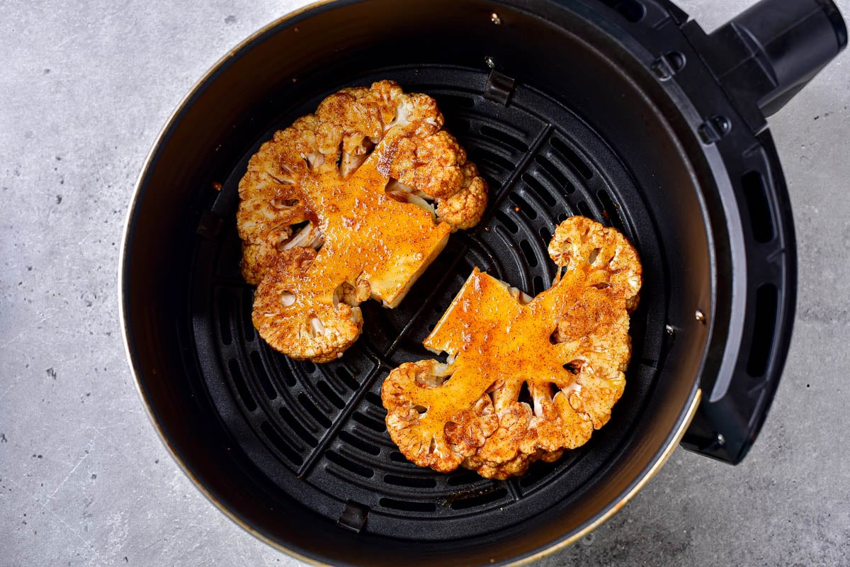 two seasoned cauliflower steaks laying flat in round black air fryer tray on counter.