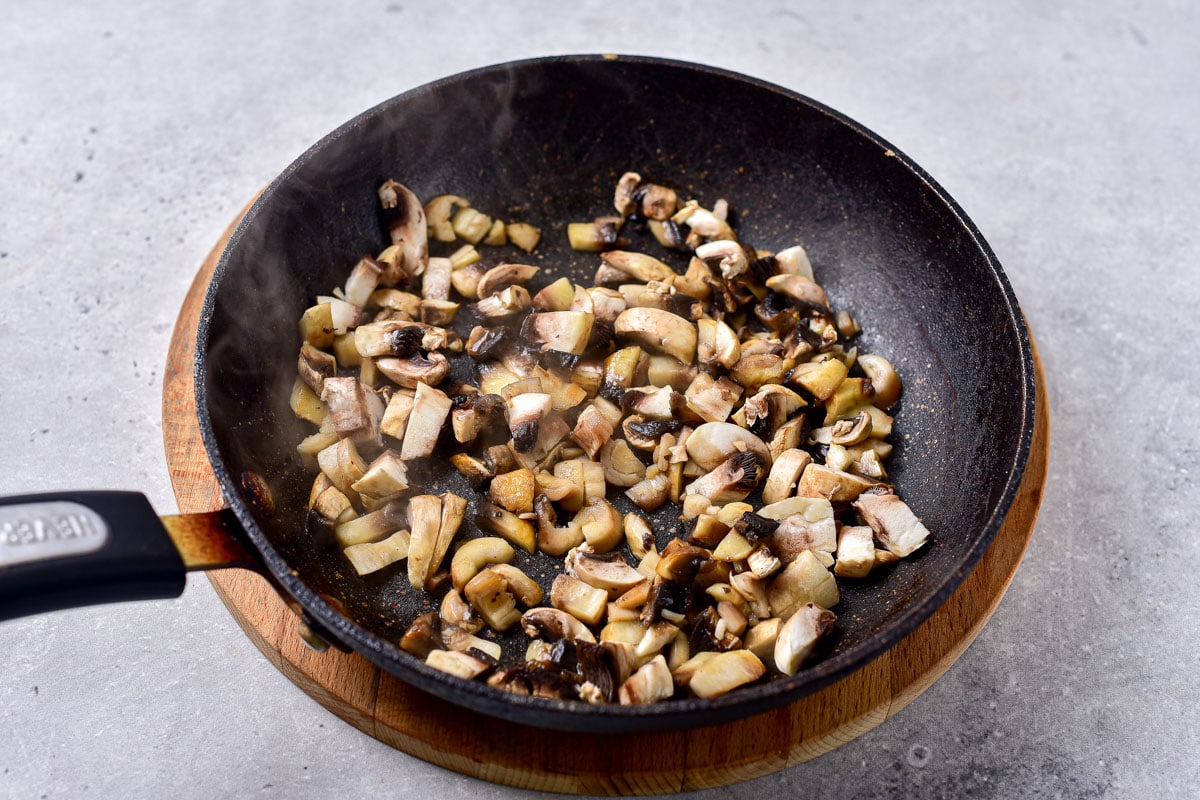 chopped mushrooms frying in black pan sitting on wood on counter.