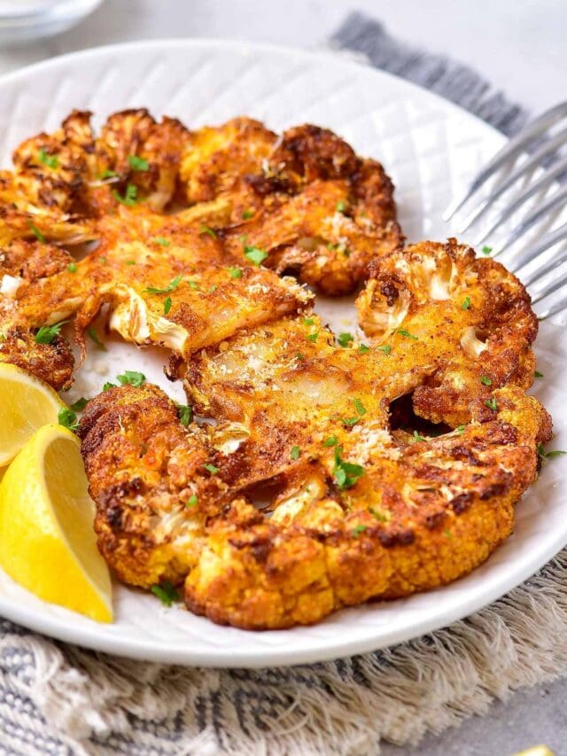 two cauliflower steaks on white plate with silver forks beside.