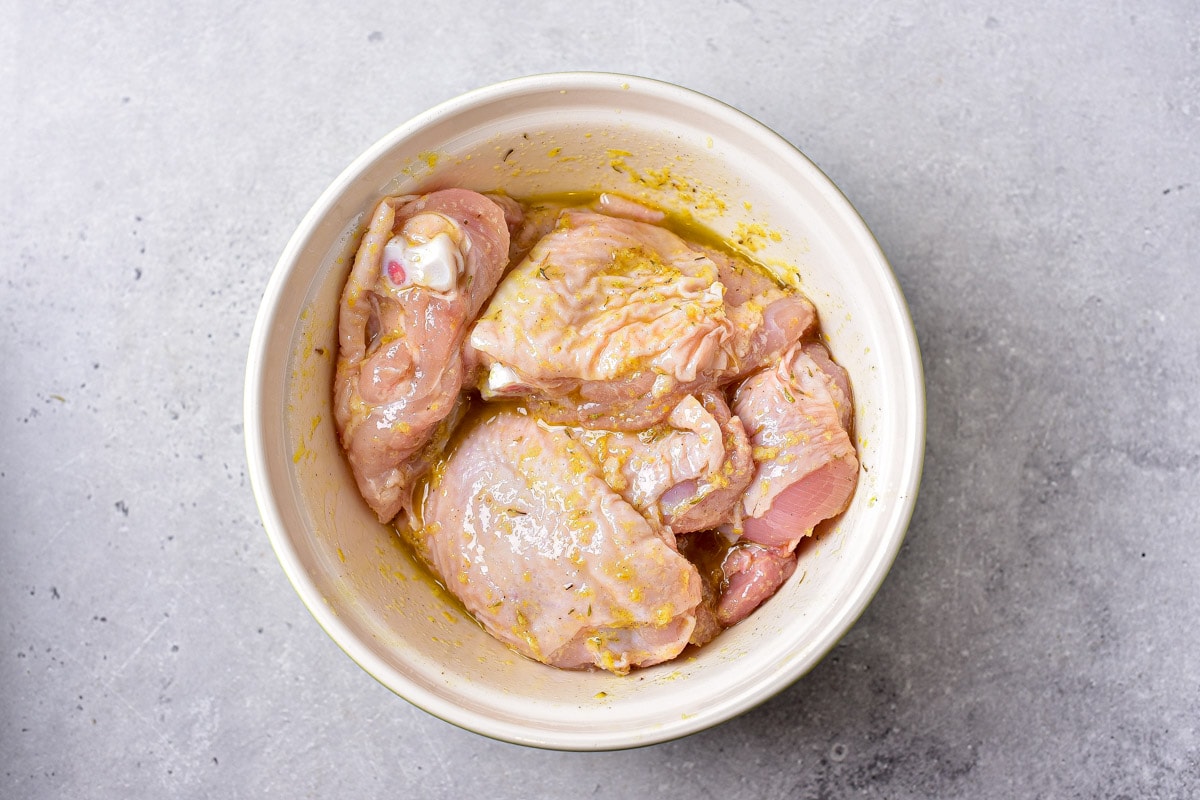 raw chicken thighs in lemon marinade in a white bowl sitting on the counter.