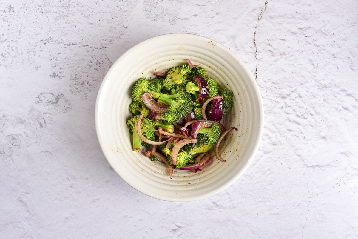 raw broccoli florets and red onion pieces in white bowl on counter.
