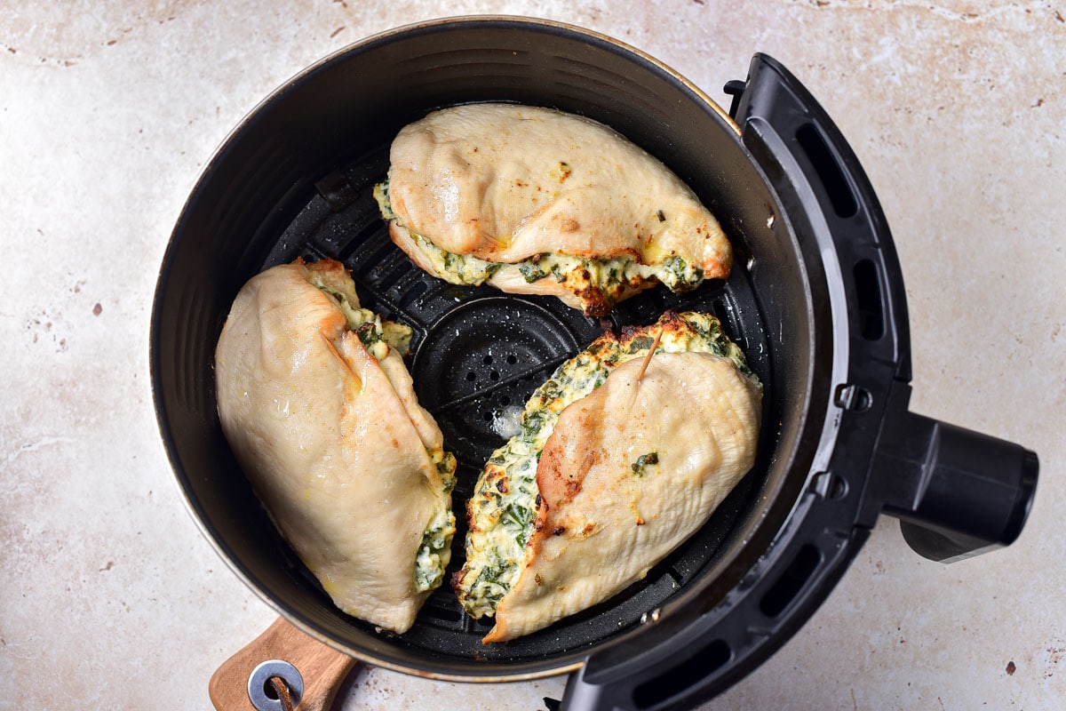 three semi-cooked stuffed chicken breasts in round black air fryer tray on counter.