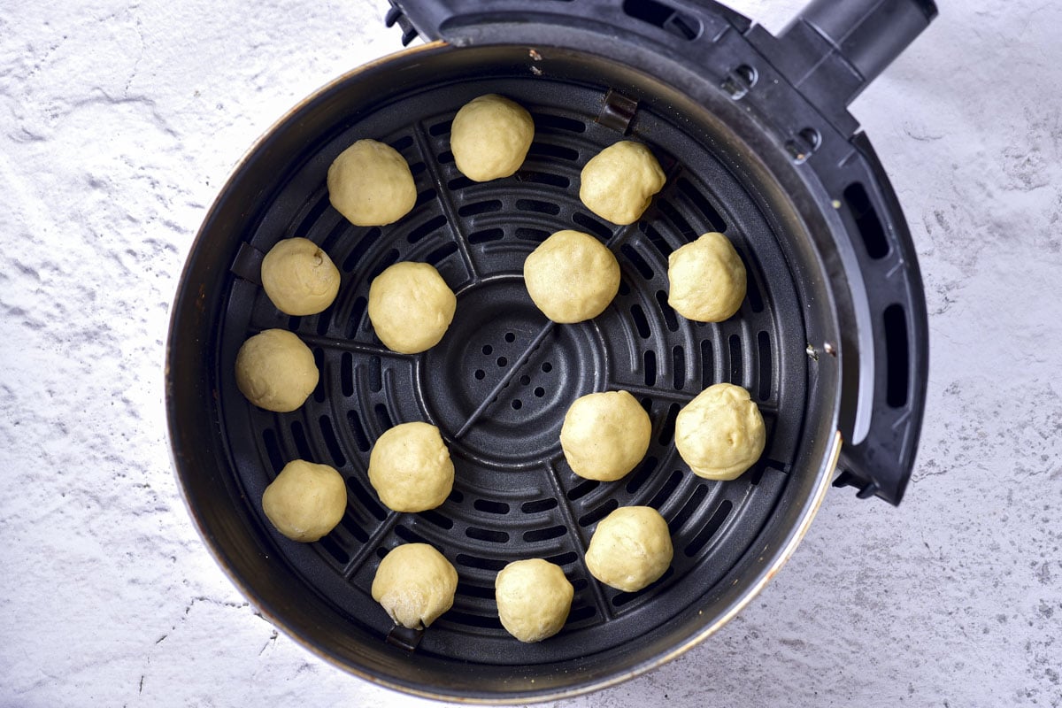 balls of raw donut dough in round black air fryer basket on counter.