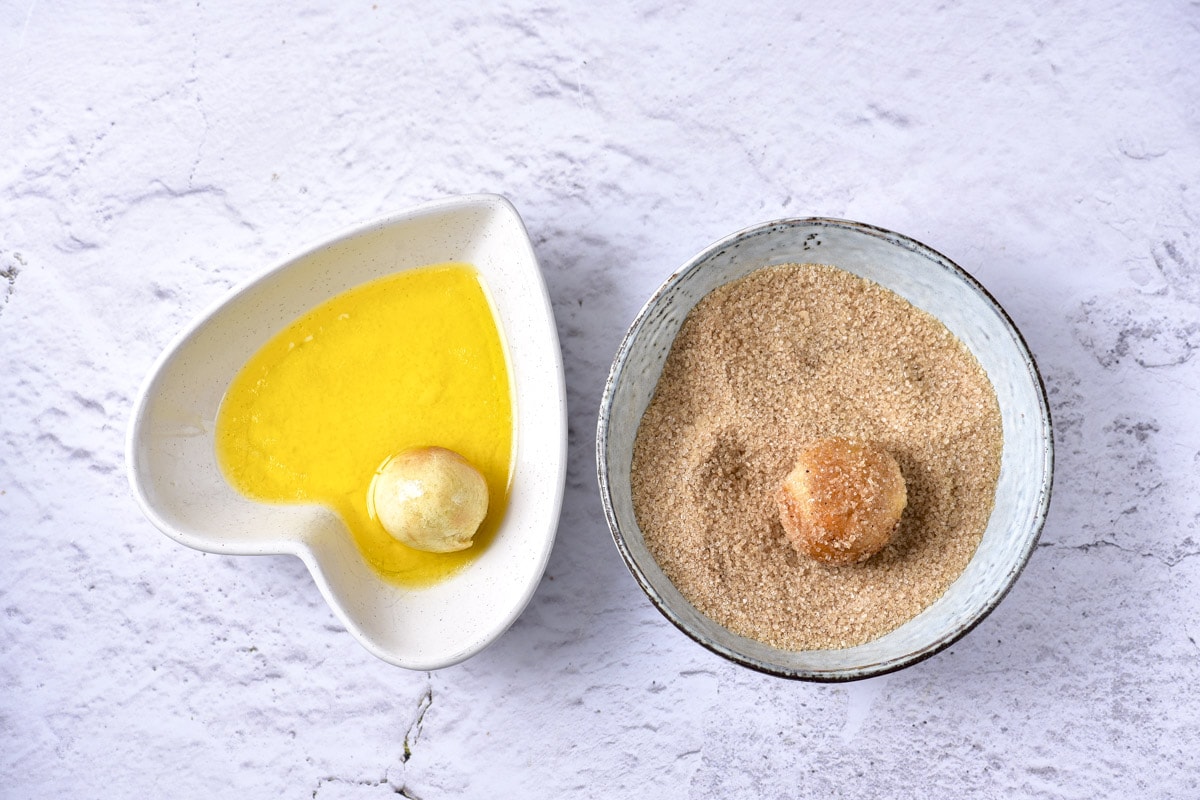 cooked donut holes being dipped in two bowls of melted butter and  cinnamon-sugar mix.