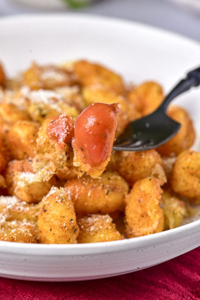 fried gnocchi dipped in sauce on the end of a fork above plate of other gnocchi.