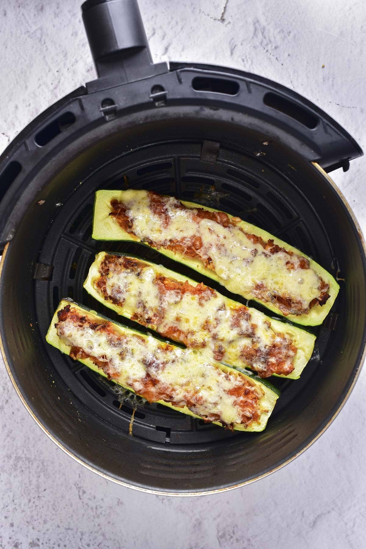cooked zucchini boats with melted cheese on top sitting in round black air fryer basket.