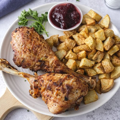 two turkey drumsticks on white plate with potatoes and cranberries beside.