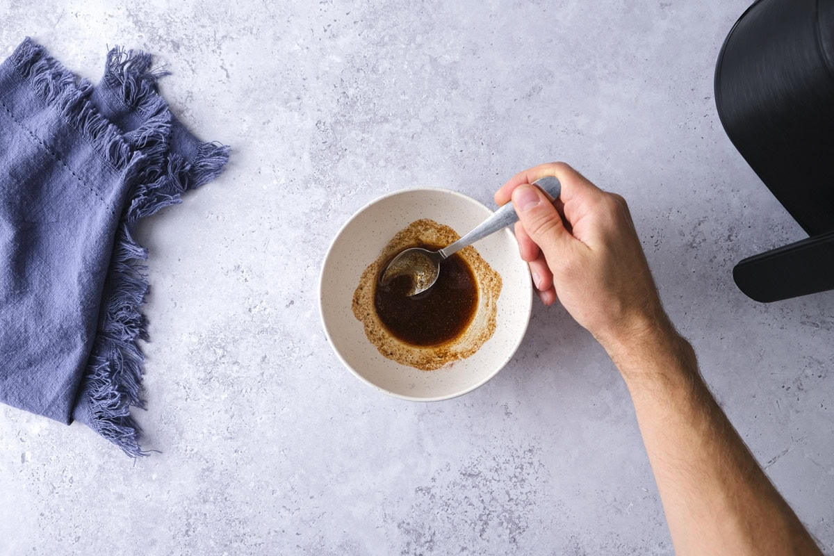 hand mixing marinade in white bowl with silver spoon on counter.