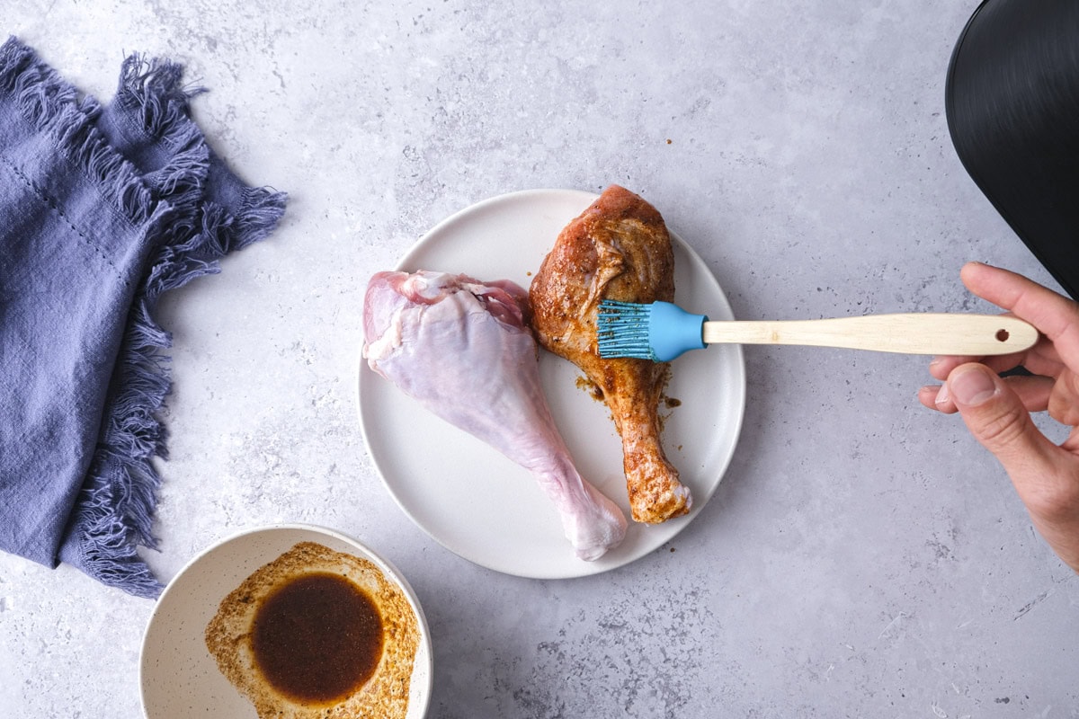 hand holding marinating brush on raw turkey drumsticks on plate.