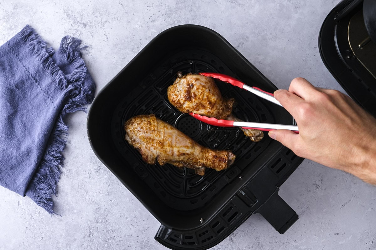red tipped tongs flipping turkey drumsticks in air fryer basket.