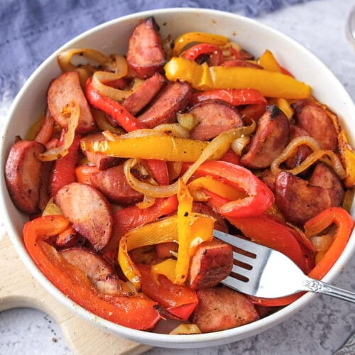 white bowl of fried kielbasa and peppers with fork sticking out.