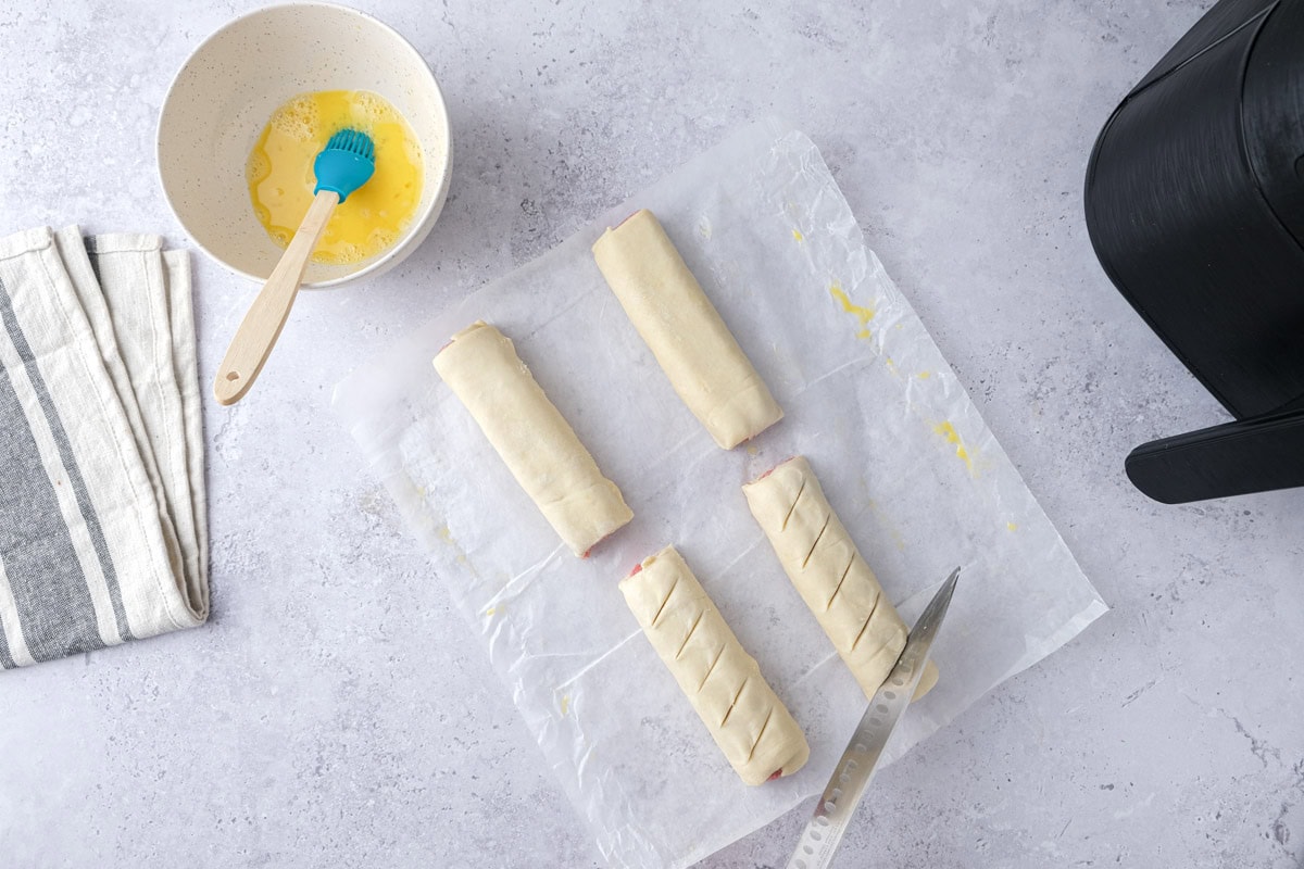 large knife cutting slits into the tops of raw sausage rolls on counter.