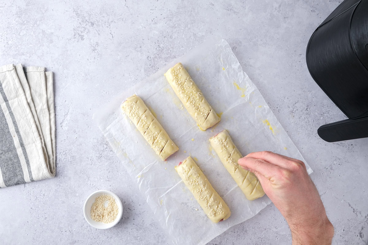hand sprinkling sesame seeds into raw sausage rolls on counter.