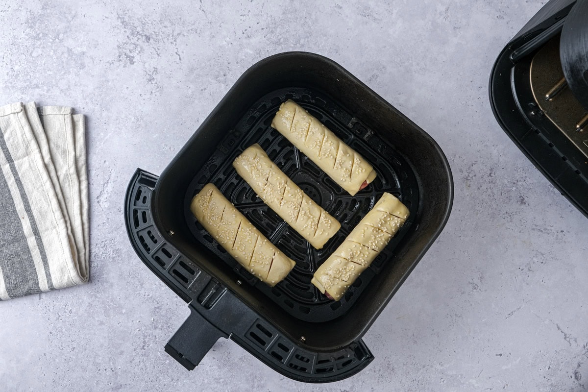 four raw sausage rolls in black air fryer tray on counter.