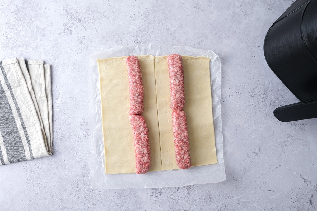 raw sausages lined up on puff pastry sheet on counter.