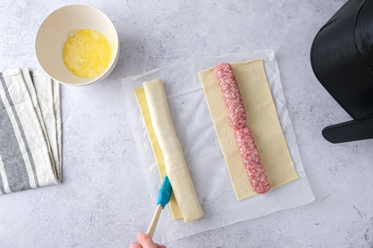 blue brush adding egg wash to inner seam of puff pastry sausage roll.