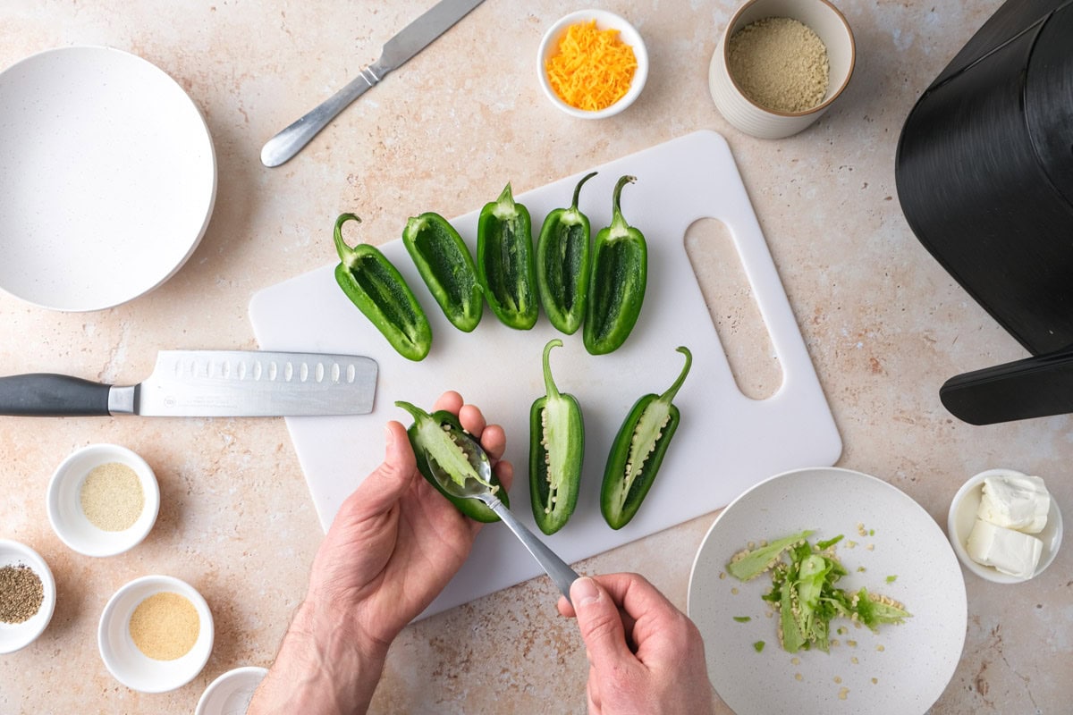 hand scooping seeds out of jalapeno peppers with counter and ingredients around.