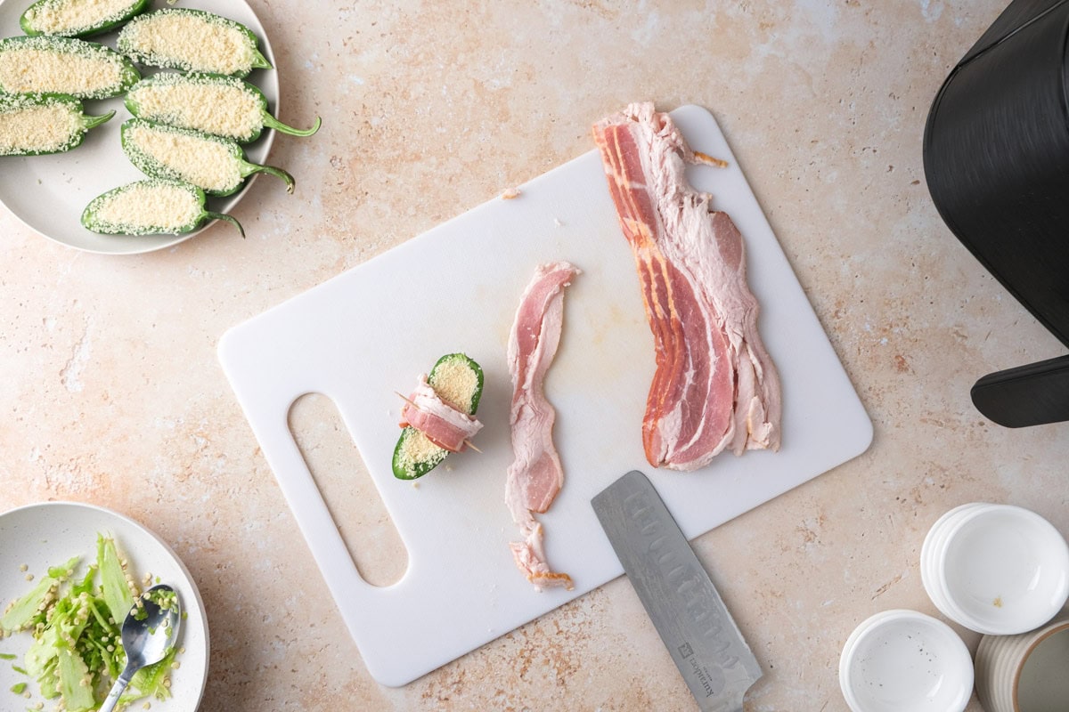 cutting board with bacon on it and one half jalapeno wrapped in one slice.