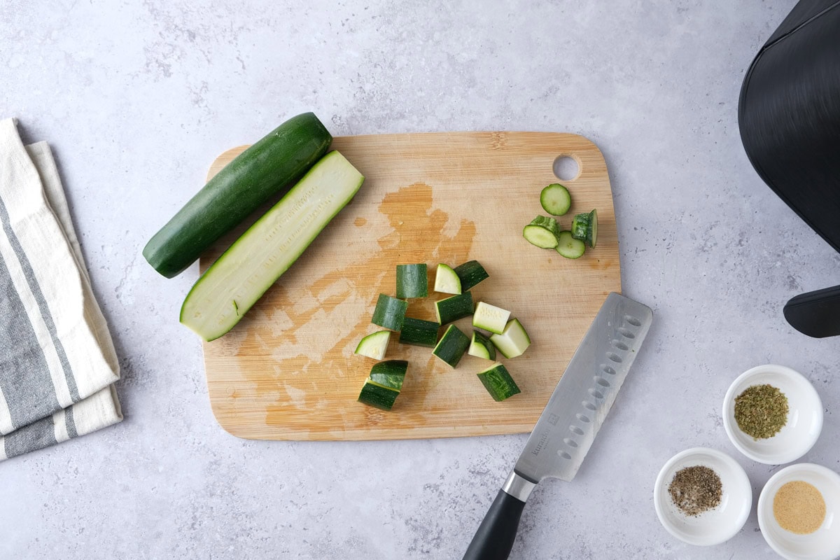 raw zucchini cut into pieces on wooden board with knife and spices beside.