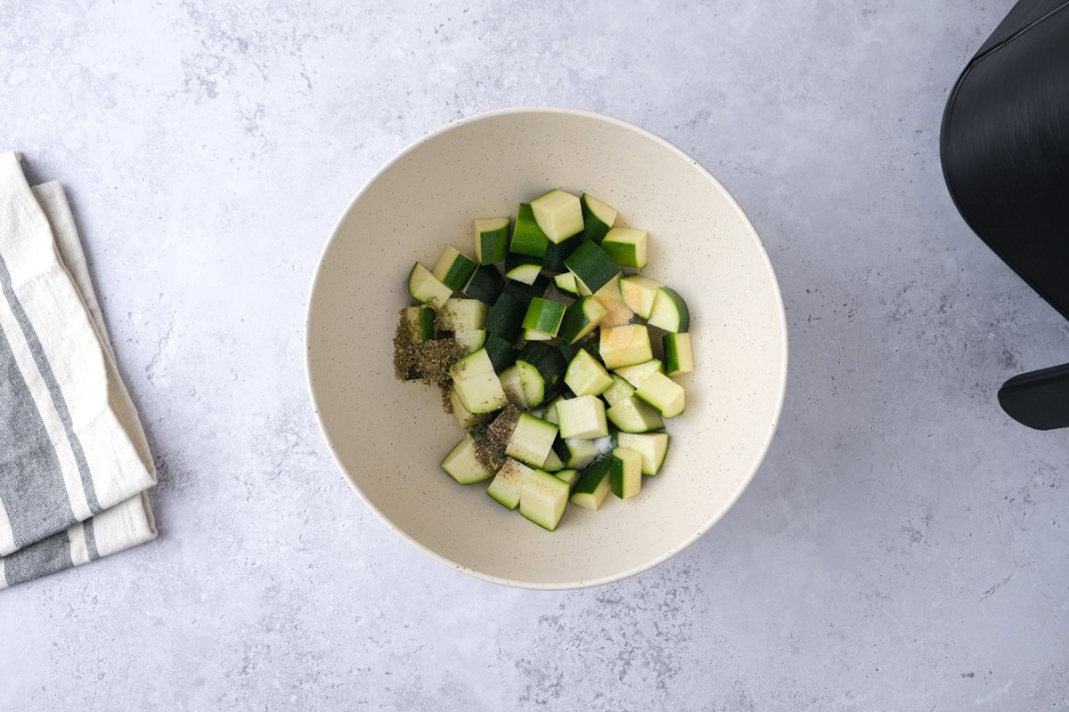 white mixing bowl on counter filled with zucchini pieces and spices.