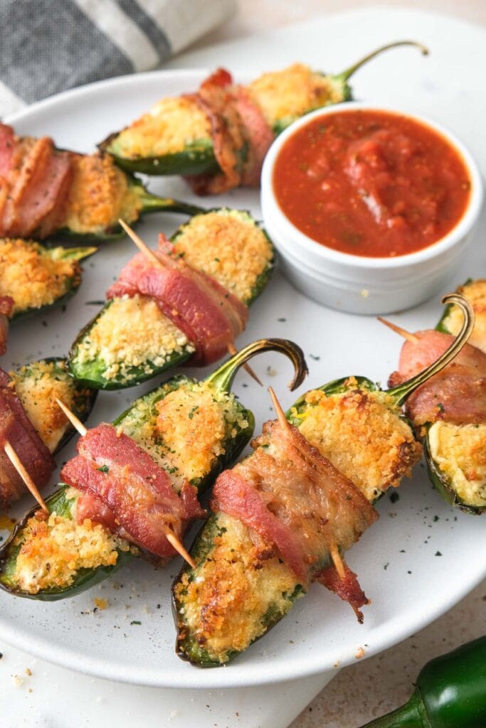 plate of crispy jalapeno poppers with red dipping sauce behind.