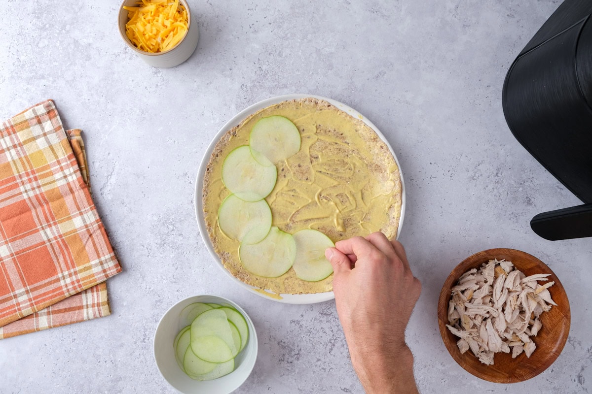 hand placing sliced green apple on a tortilla covered in mustard.