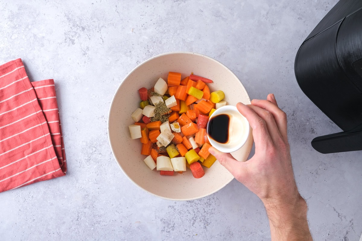 hand pouring balsamic vinegar into bowl of cut root vegetables.