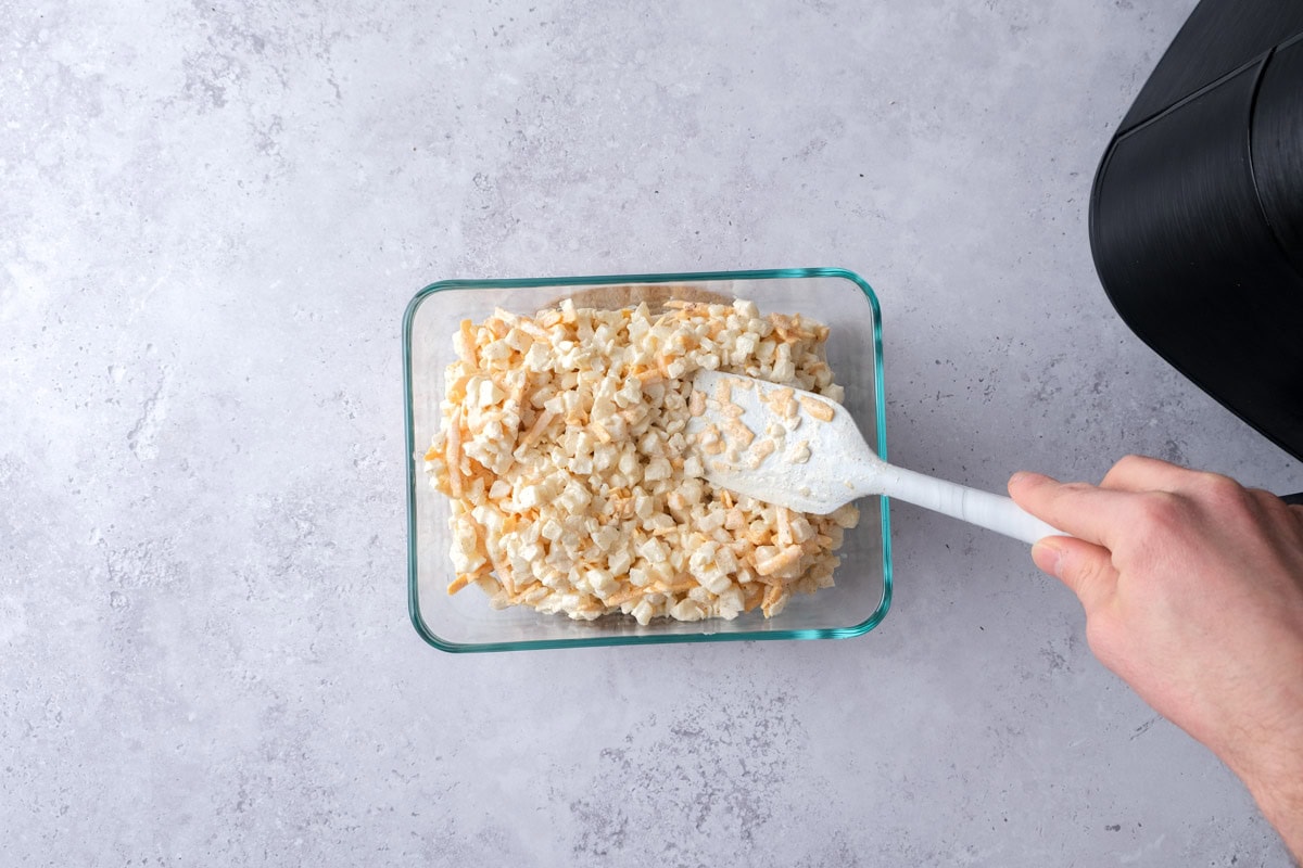 spatula spreading out raw potato cheese mixture in glass dish on counter.