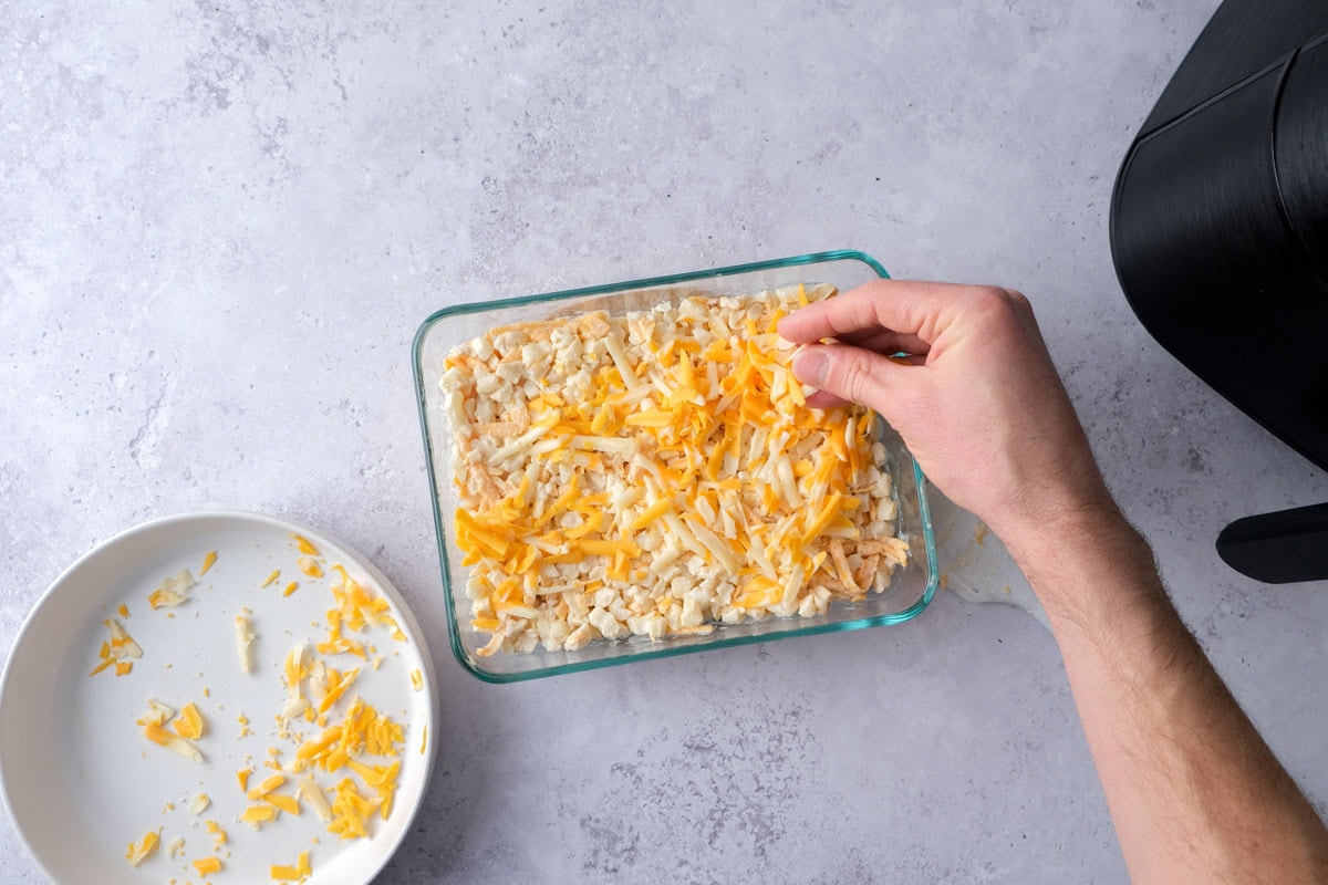 hand adding grated cheese to the top of a potato bake in glass dish.