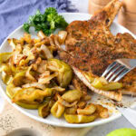 plate of pork chops and apples on wooden board with fork and knife around.