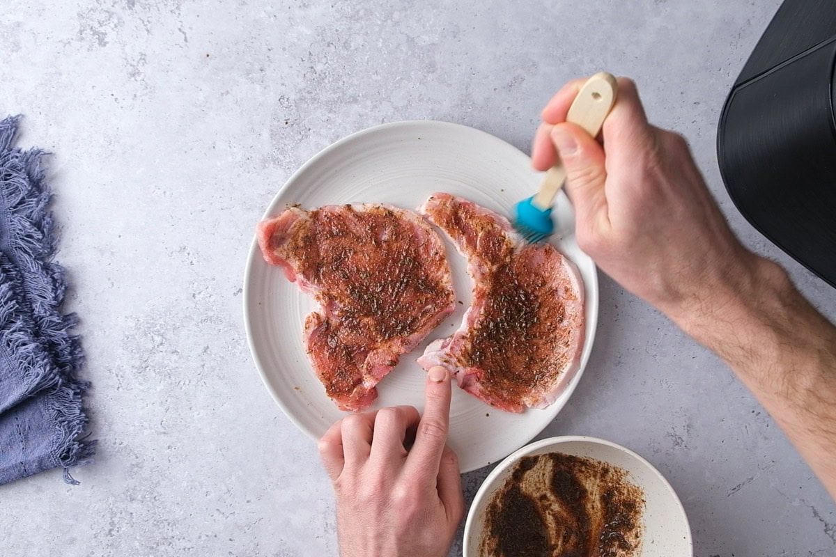 hand using brush to marinate raw pork chops on plate.