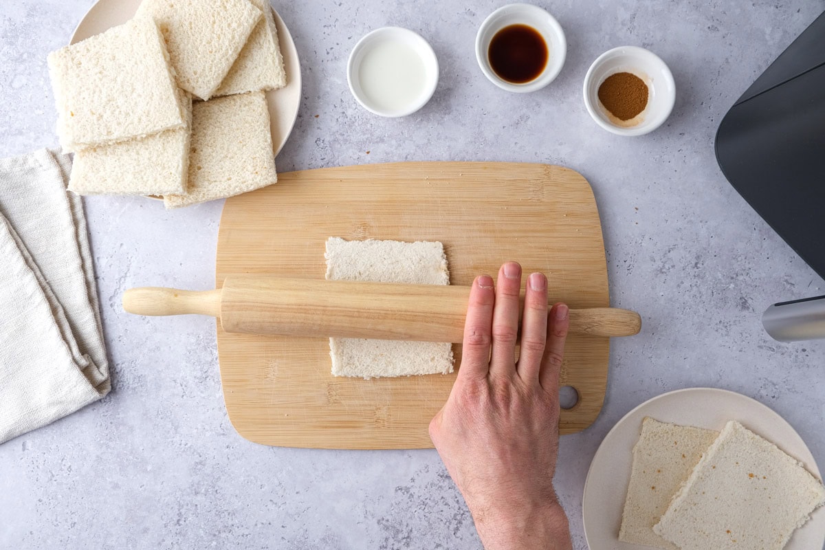 hand rolling out squares of white bread on wooden cutting board.