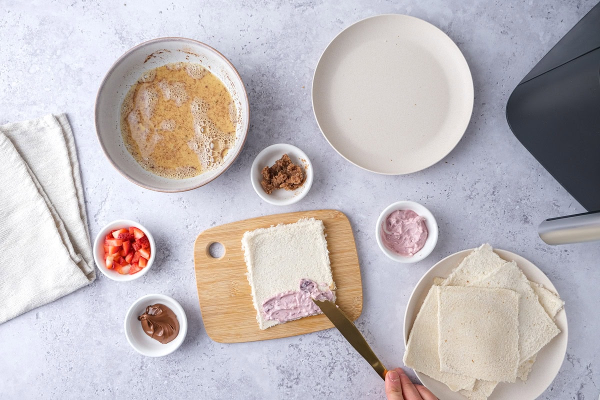 knife spreading jam and cream cheese on square of bread with ingredients around.