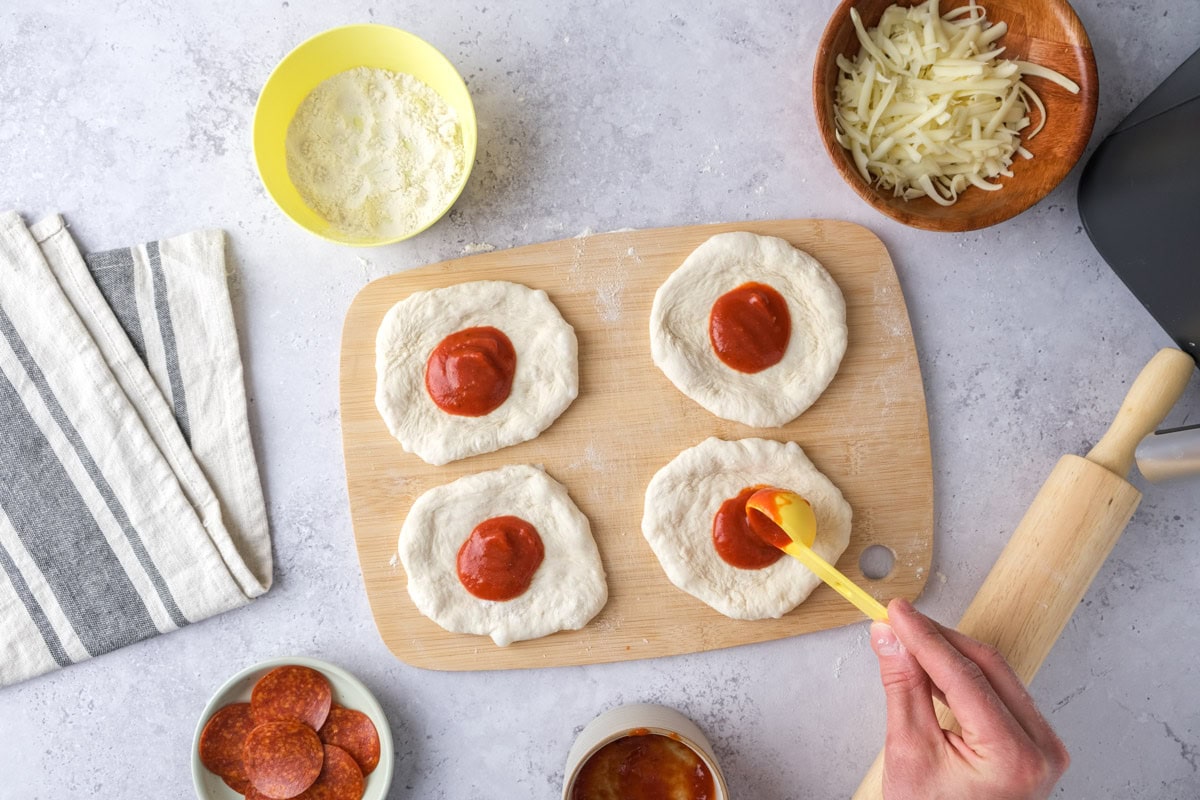 hand using spoon to add pizza sauce to dough circles on counter.
