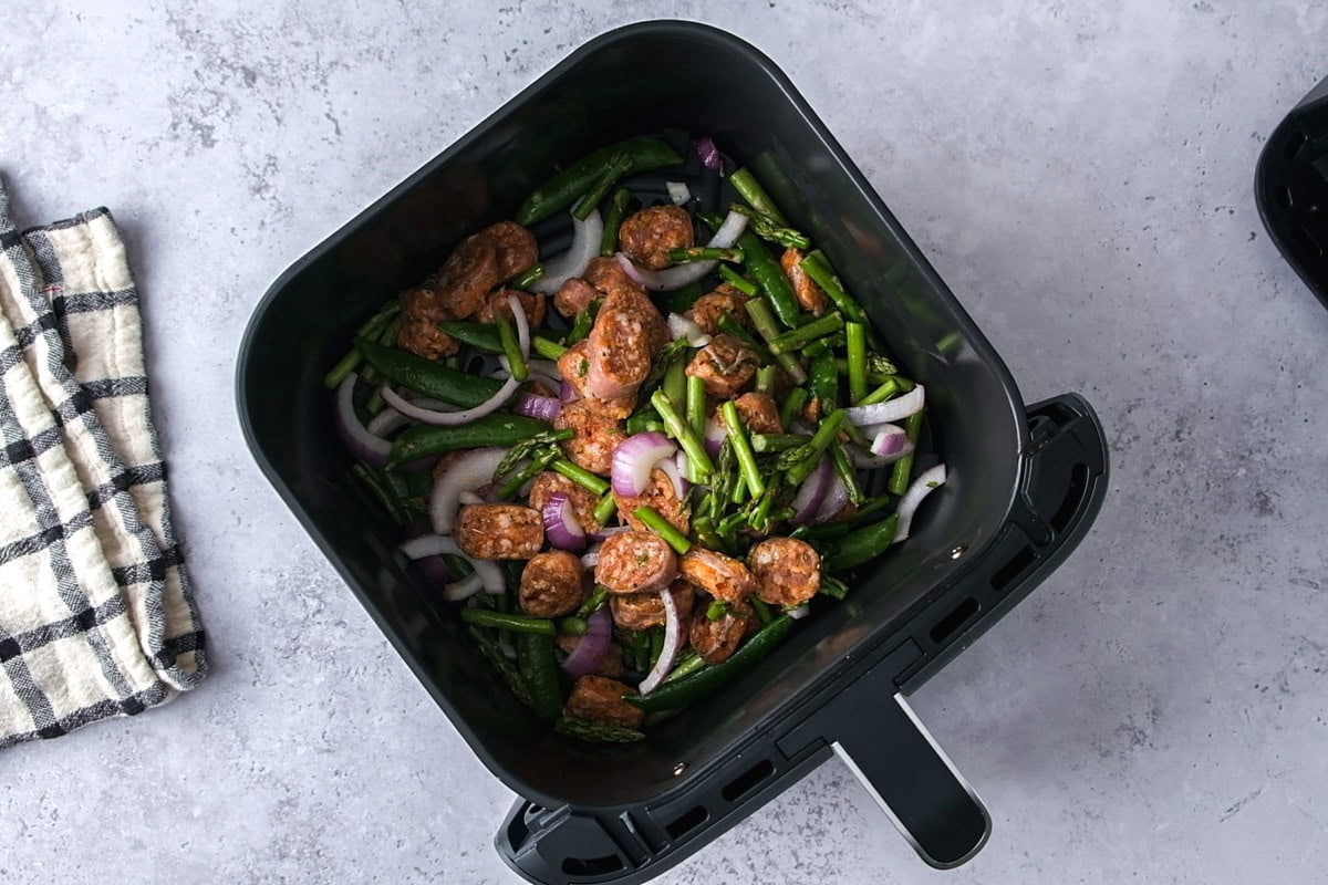 raw sausage and vegetables in black air fryer basket on counter.