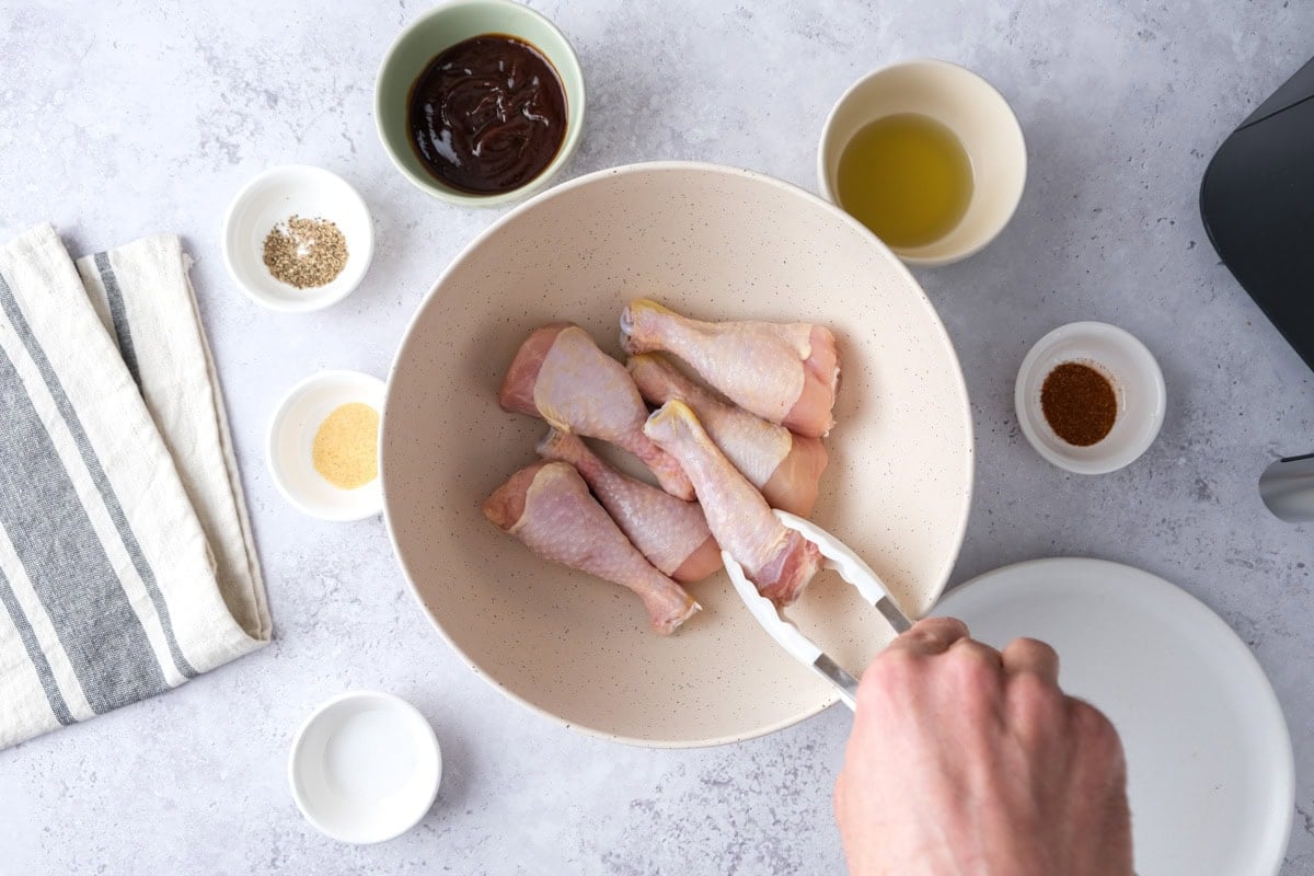 hand using tongs to place raw chicken drumsticks into a mixing bowl on counter.