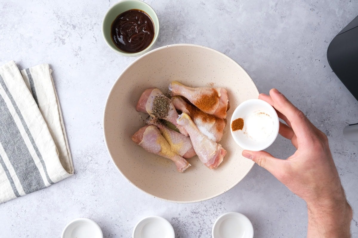 hand pouring spices into bowl of raw chicken drumsticks on counter.